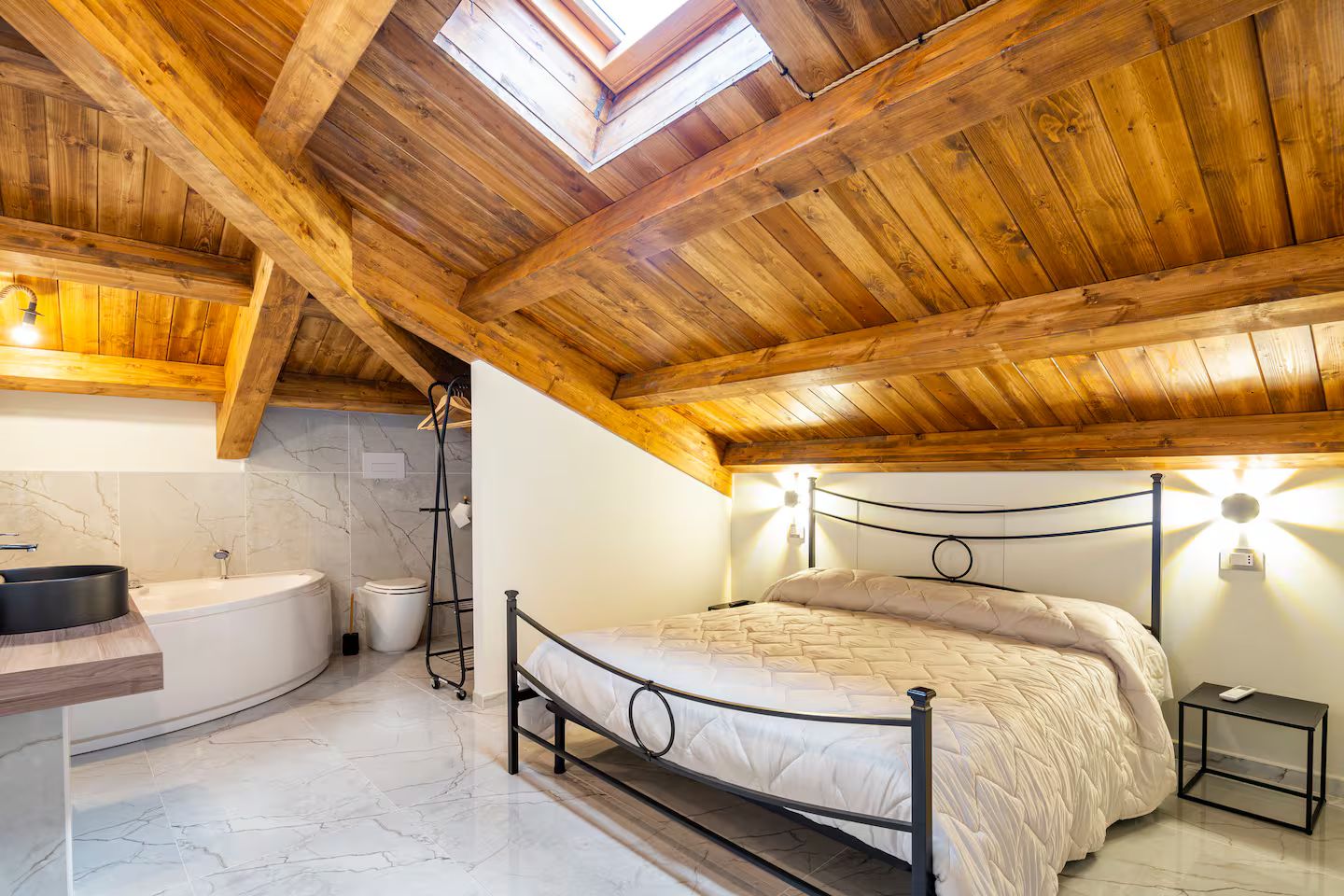 A bedroom with a wooden ceiling, a bed, a nightstand, a bedside table, and a skylight.