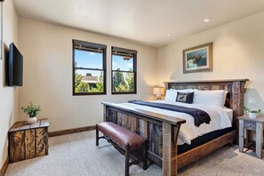 A bedroom with a large bed, a bench, a nightstand, a wall-mounted TV, and two windows. - Spacious 6700 SQFT Home | 4 Ensuite Bedrms + Kids Bunkrm + Game Rm + Media Room (Powell Butte)