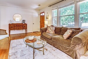 A living room with a brown sofa, a glass coffee table, a wooden dresser, and a large window with a view of trees. - 9 Mi to Dtwn Nashville! Updated Home + Large Yard (Nashville)