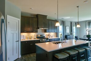 A modern kitchen with dark cabinetry, a central island with bar stools, and pendant lighting. - Stylish Waterfront Home w/ Views in Lewes! (Lewes)
