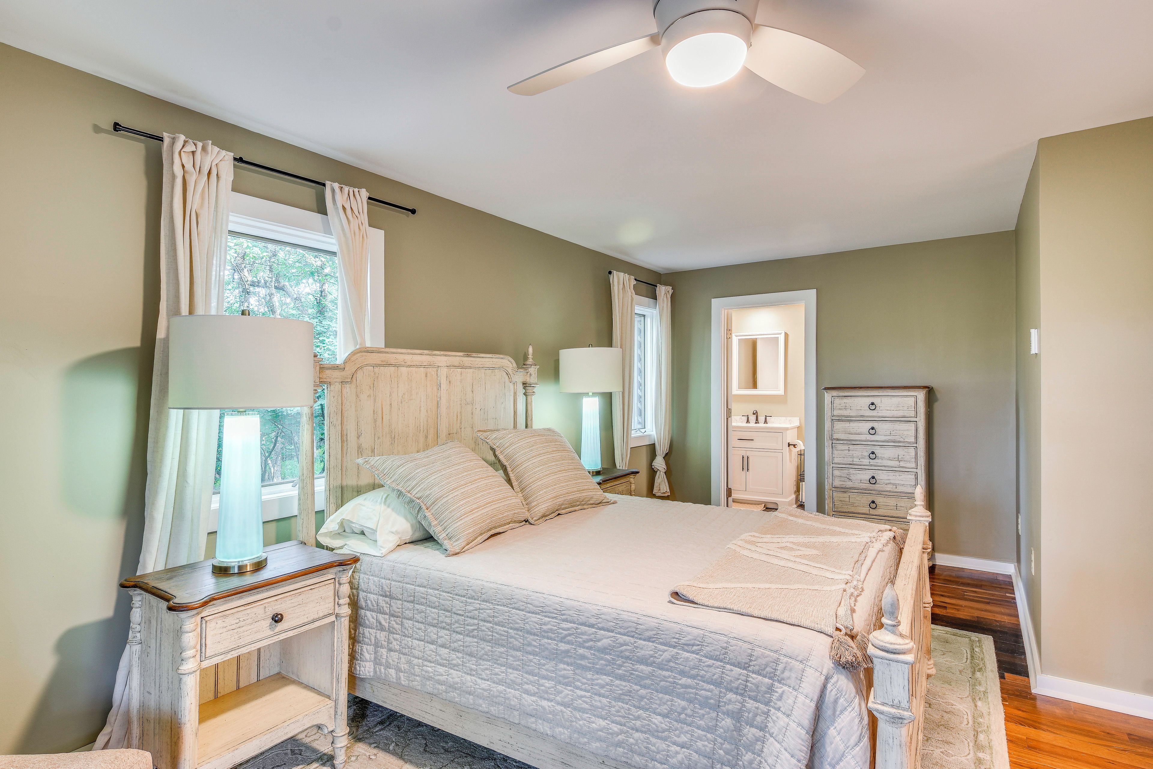 Une chambre à coucher comprenant un lit, des tables de chevet, une commode, un ventilateur de plafond et une fenêtre avec des rideaux.