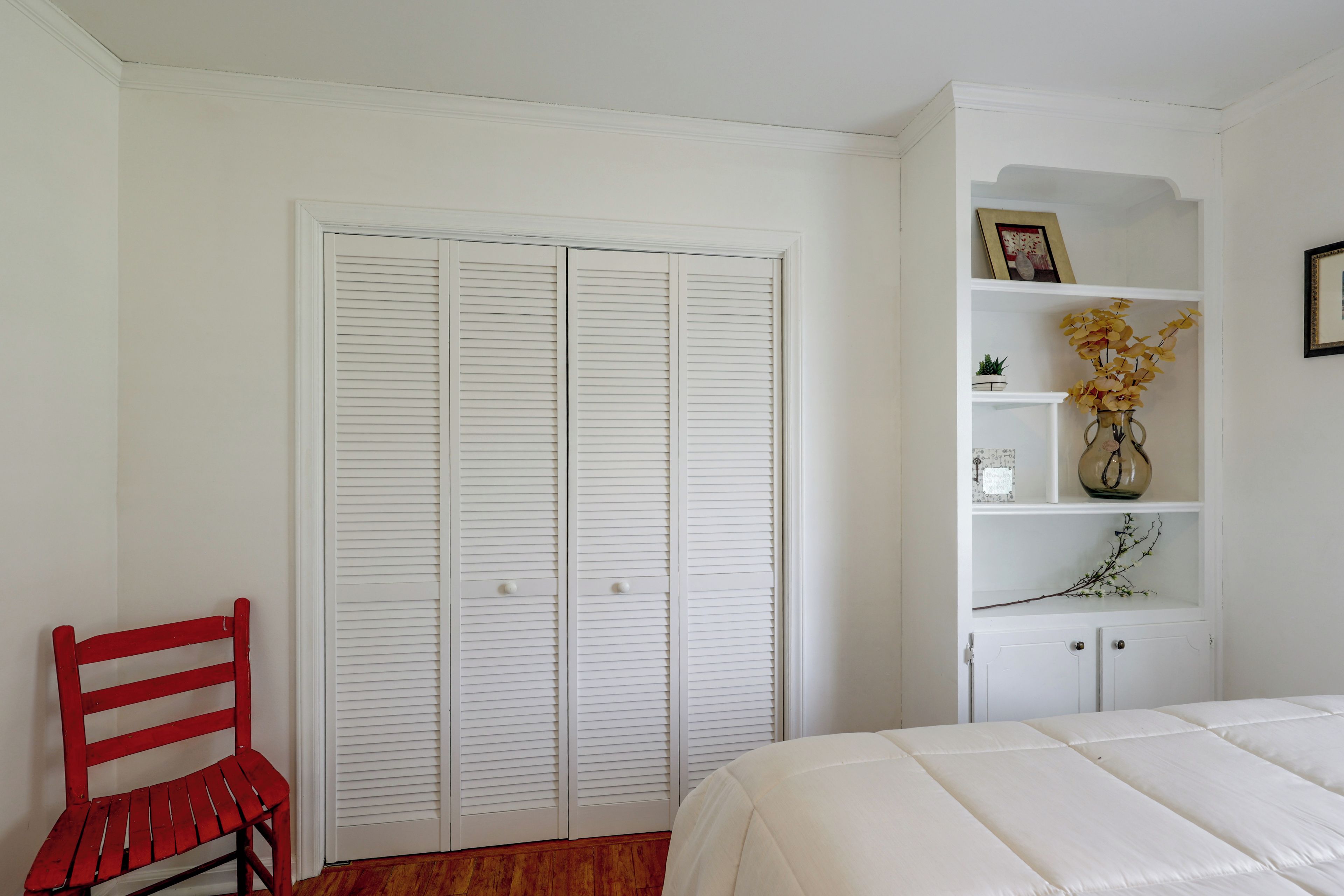 A bedroom with a white bed, a red chair, a white wardrobe, and a shelf with decorative items.
