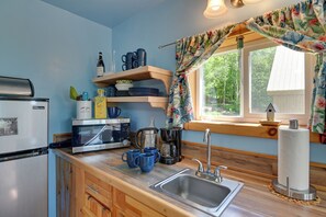 A compact kitchen with a refrigerator, microwave, coffee maker, and a sink. There are wooden shelves above the counter holding dishes and a birdhouse on the window sill.