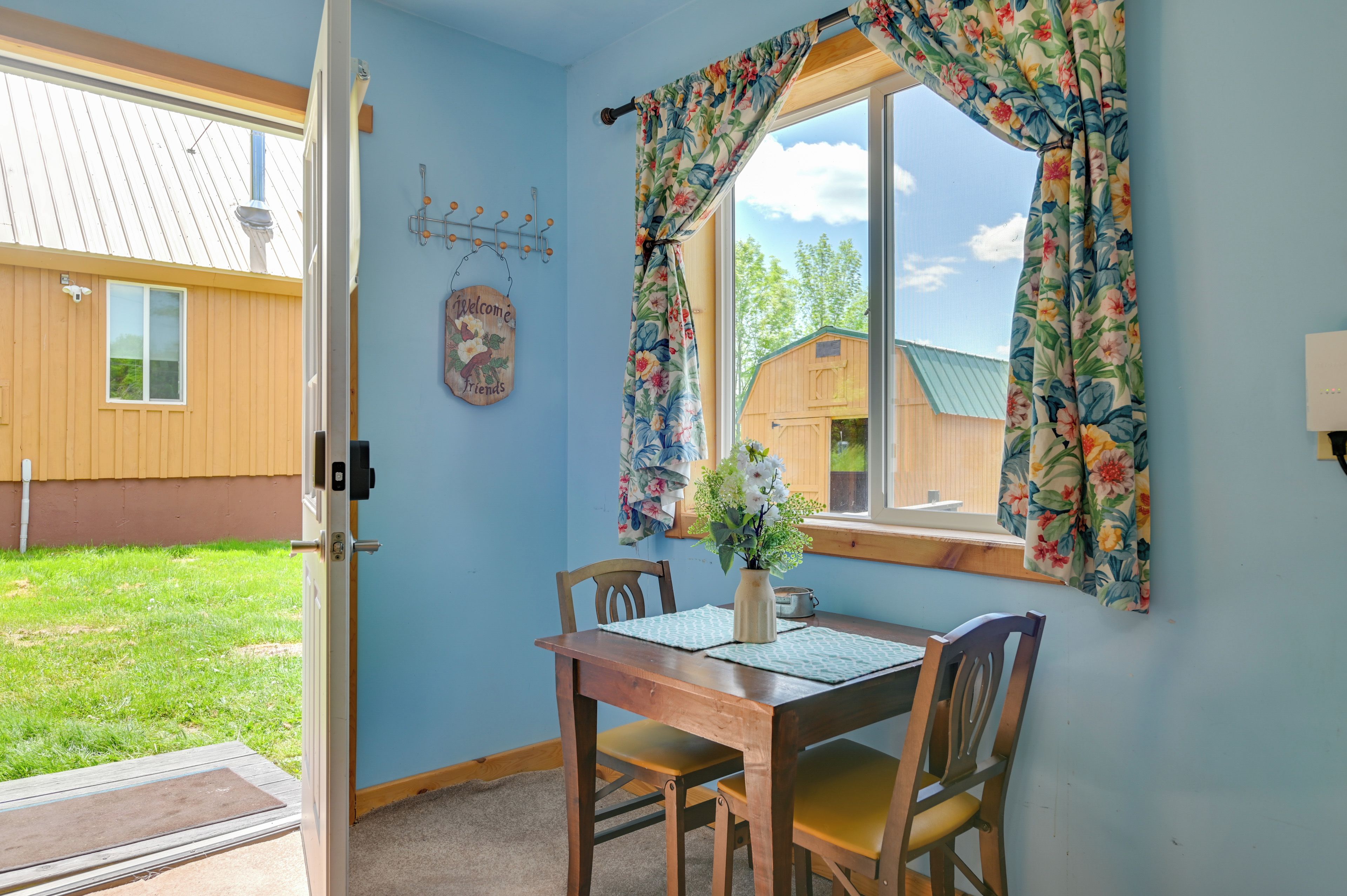 A room with a wooden table and chairs, a window with floral curtains, a welcome sign, and a view of a wooden building outside.