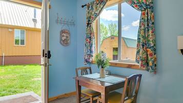 A room with a wooden table and chairs, a window with floral curtains, a welcome sign, and a view of a wooden building outside.