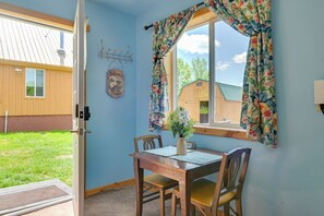 A room with a wooden table and chairs, a window with floral curtains, a welcome sign, and a view of a wooden building outside.