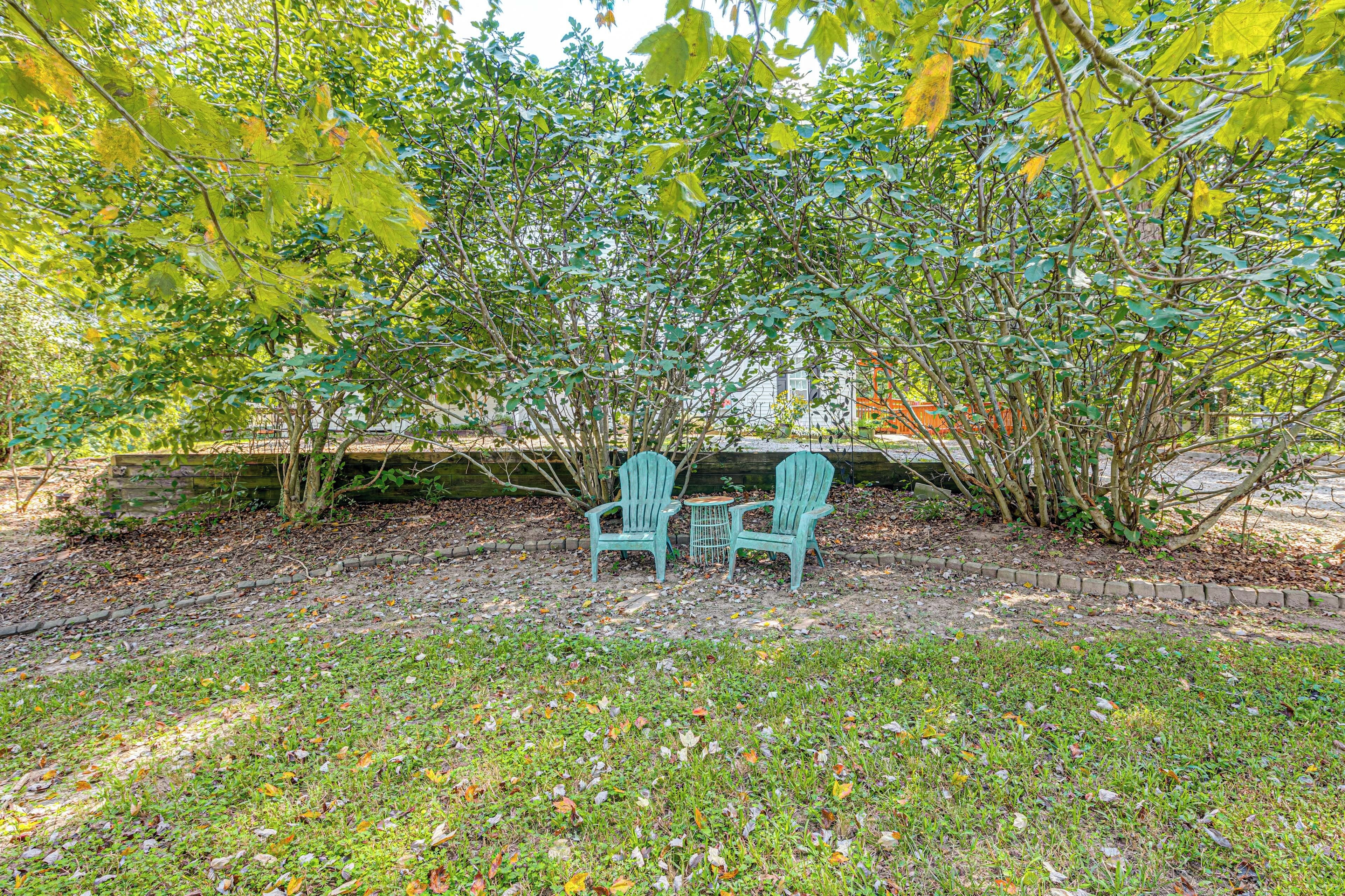 A garden area with two Adirondack chairs and a small table, surrounded by trees and a brick wall.
