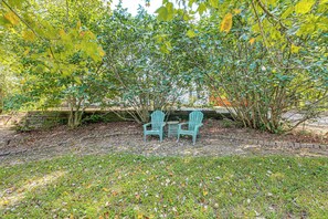 A garden area with two Adirondack chairs and a small table, surrounded by trees and a brick wall.