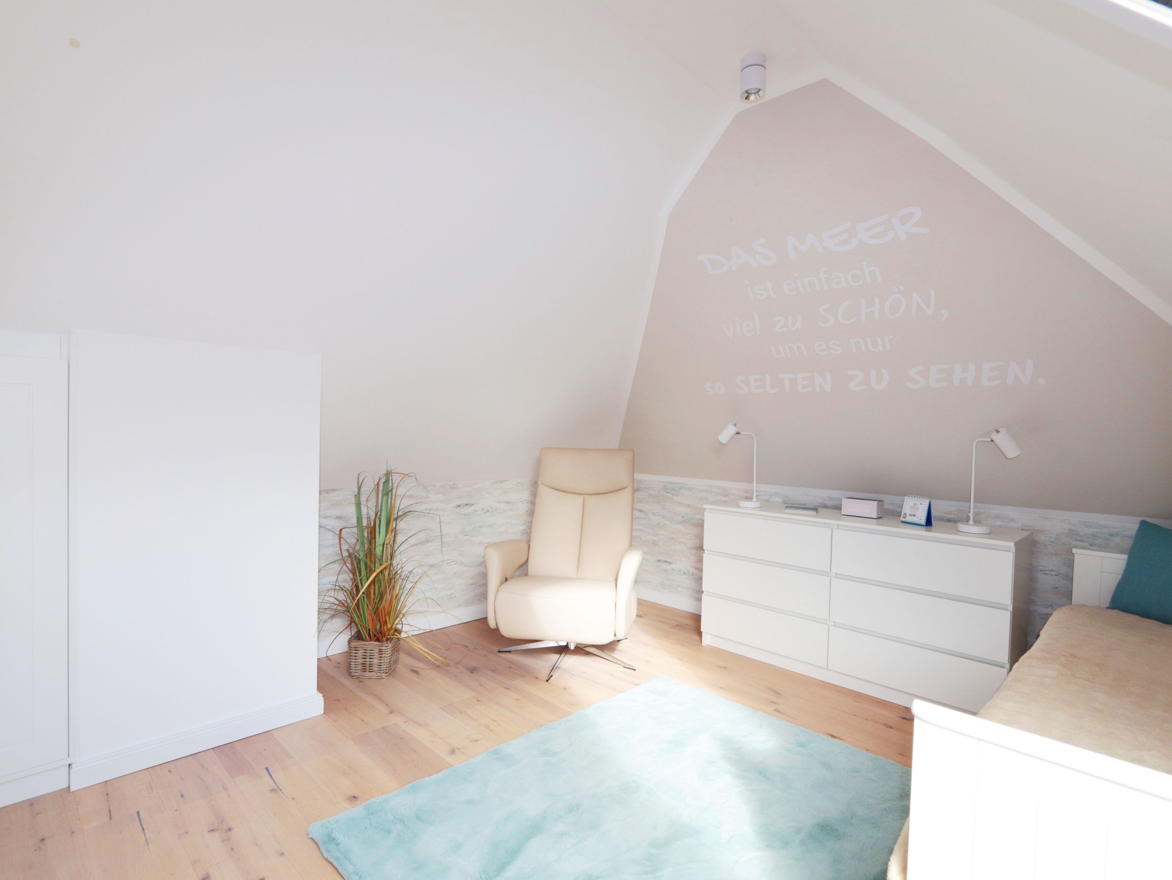 Una habitación con techo inclinado, una silla blanca, un tocador blanco, una planta pequeña y una alfombra azul claro.
