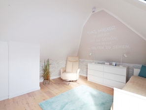 A room with a sloped ceiling, a white chair, a white dresser, a small plant, and a light blue rug. - Holiday Apartment 'Kleine Meerzeit Zingst' with Gas Fireplace, Underfloor Heating, Balcony, Wi-Fi, Central & Quiet (Zingst)