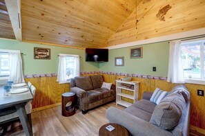 A cozy cabin interior with a wooden ceiling, a television mounted on the wall, and a dining area with a table set for two. - Walk to Lake Ontario! 'Minnow Cottage' (Kent)