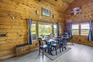 A wooden cabin interior with a dining table set for four, a bench, and a view of the outdoors through large windows.