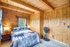 A wooden cabin bedroom with a bed, two bedside lamps, a window with curtains, and a standing fan.