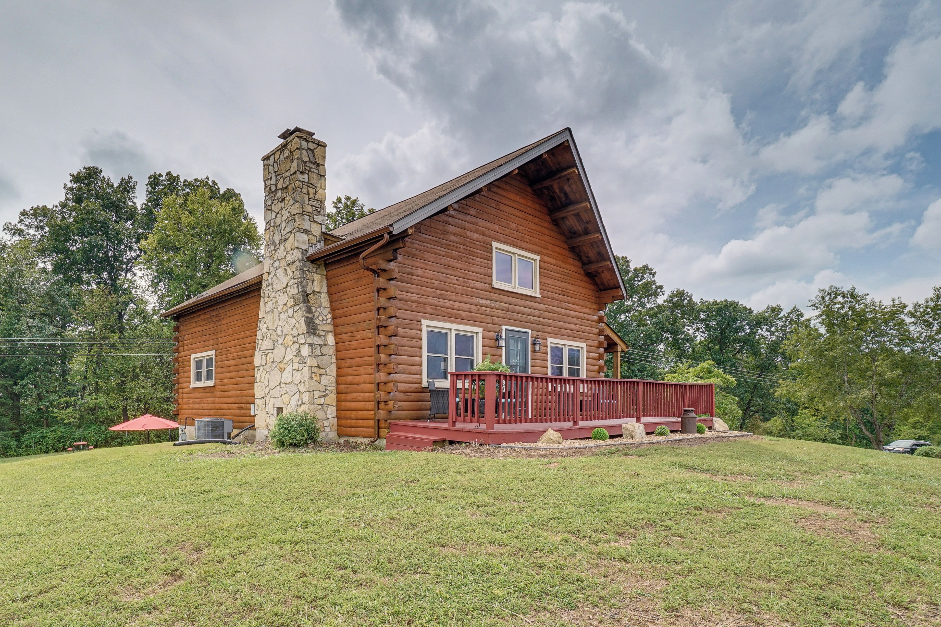 A log cabin with a stone chimney, a red deck, and a grassy yard.