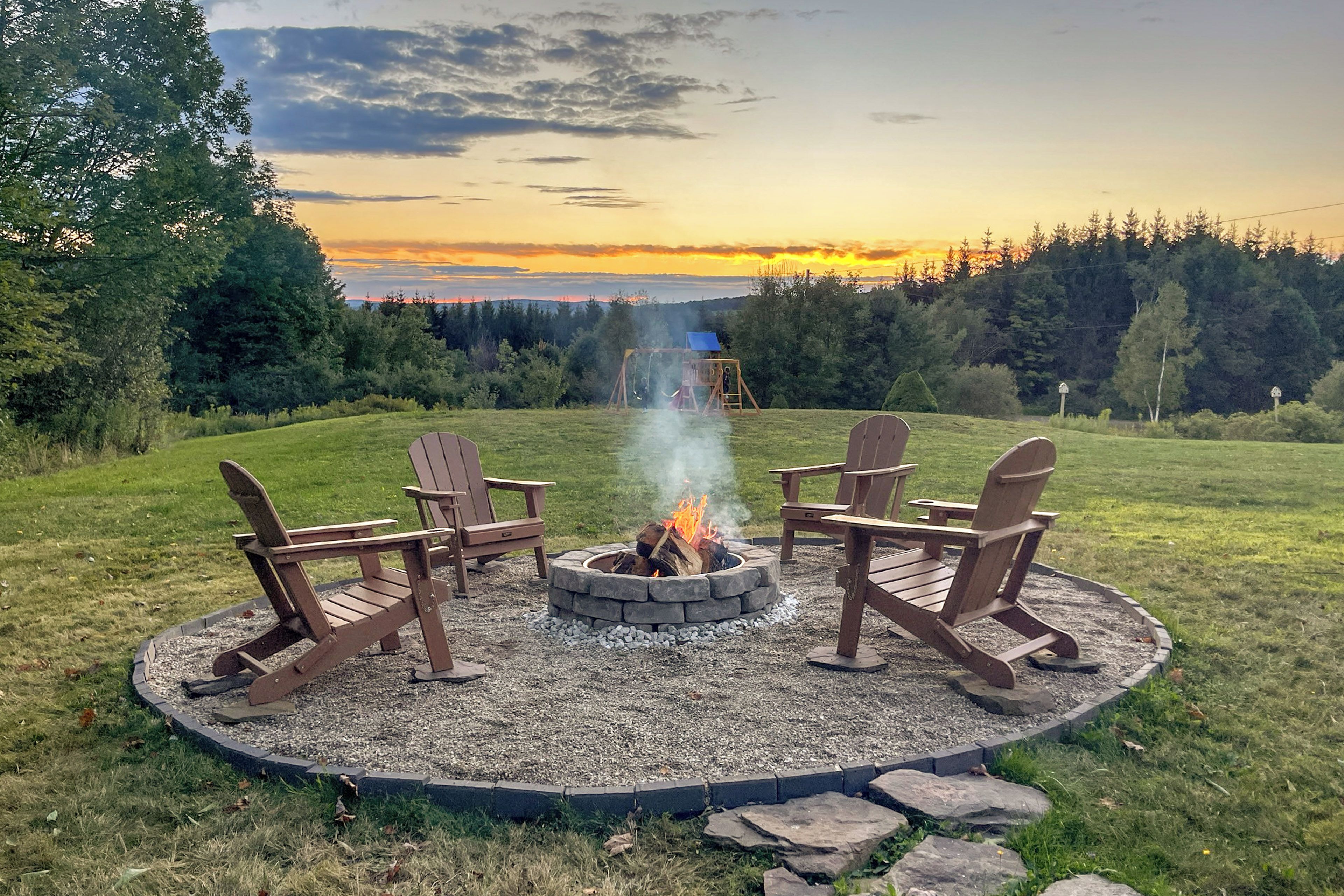 A circular fire pit with four Adirondack chairs around it, set in a grassy area with a view of a forested landscape and a sunset in the background.