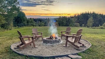 A circular fire pit with four Adirondack chairs around it, set in a grassy area with a view of a forested landscape and a sunset in the background.