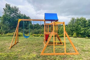 A wooden swing set with a blue roof and a red slide in a grassy area with trees in the background.