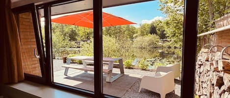 A room with large windows overlooking a garden with a pond, picnic tables, and an outdoor umbrella.