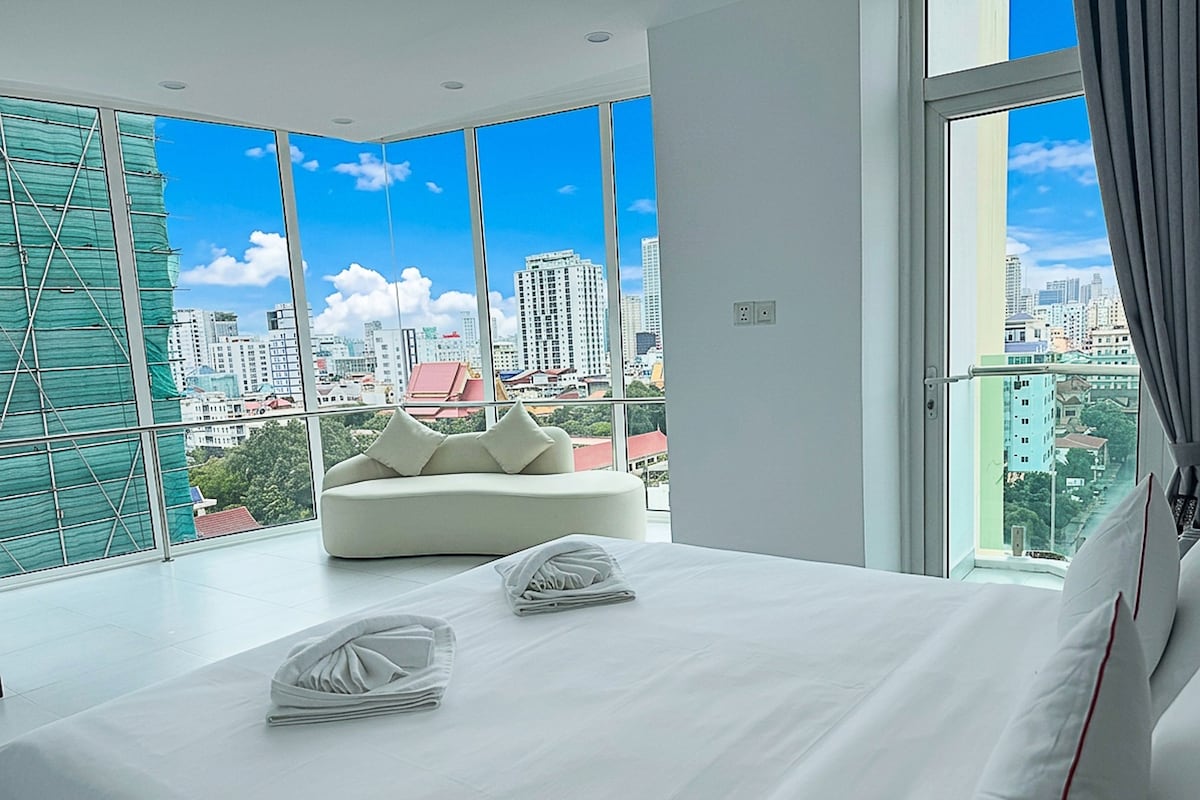 Ein modernes Hotelzimmer mit einem großen Bett, einer Couch und Blick auf die Stadt durch raumhohe Fenster.
