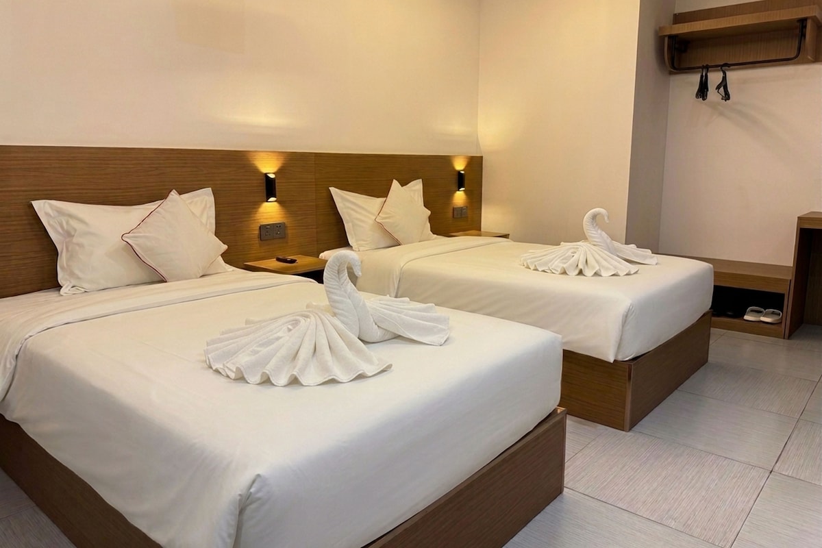 Ein Hotelzimmer mit zwei Betten, jeweils mit ordentlich gefalteten Handtüchern und einer Schwanenform. Über den Betten befinden sich Wandleuchten und rechts daneben ein Schuhregal.
