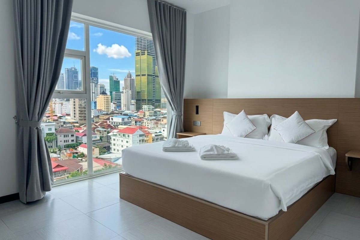 Ein Hotelzimmer mit einem großen Bett, einem Nachttisch und Blick auf eine Stadtkulisse durch ein großes Fenster.