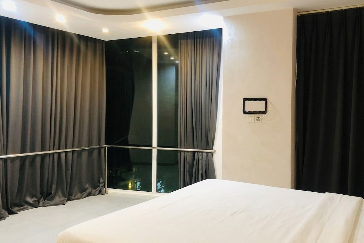 Ein Hotelzimmer mit einem großen Bett, einem Vorhangfenster, einer Bedienkonsole an der Wand und Blick ins Freie.