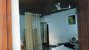 A bedroom with a bed, a bedside table, a chair, a curtain, and a ceiling fan. - Tulip Garden And Resort (Bichhiya)