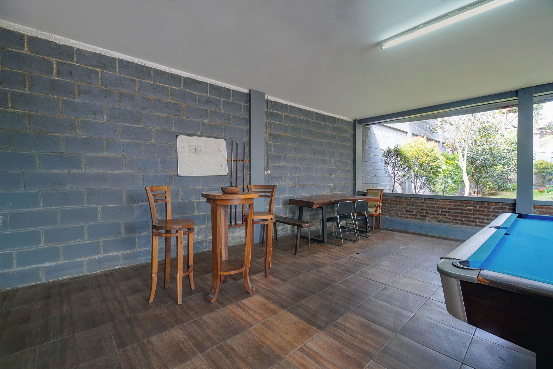 Una mesa de billar en una habitación con pared de ladrillo y ventana con vistas a un jardín.