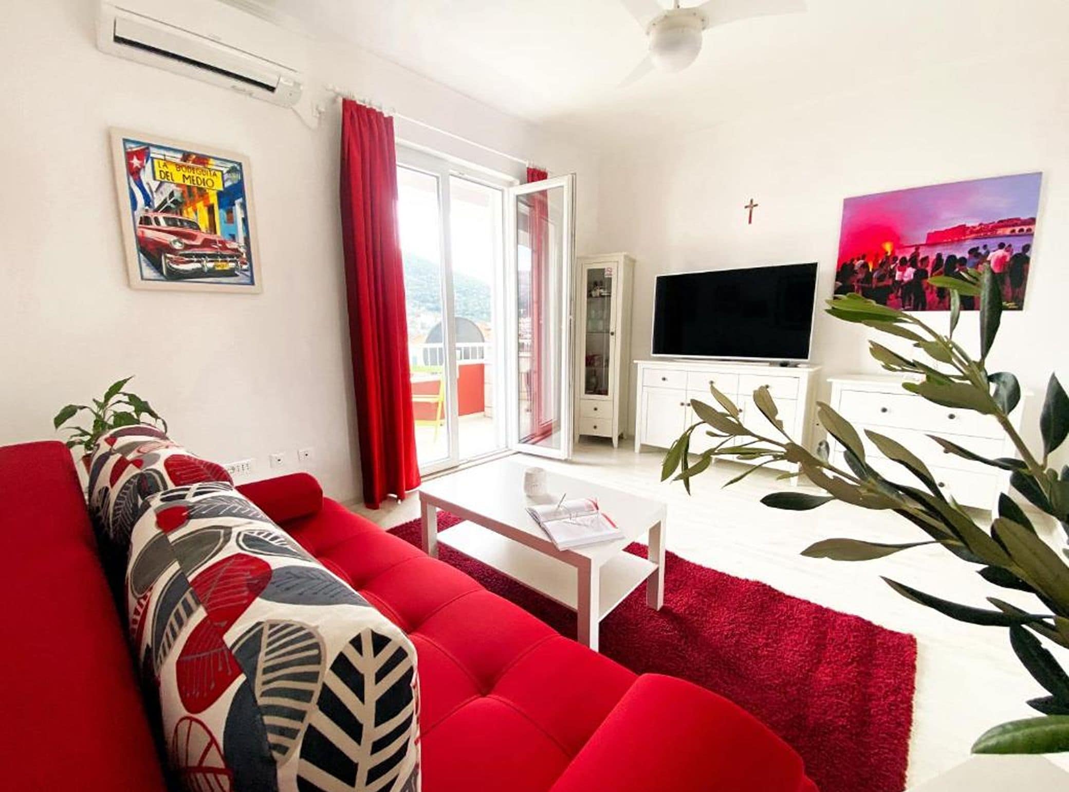 Una sala de estar con un sofá rojo, una mesa de centro blanca y un televisor sobre un mueble blanco.
