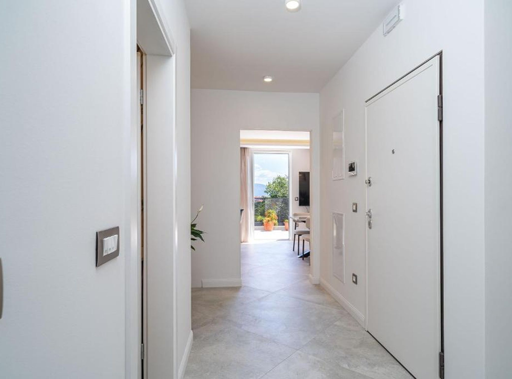 A hallway with a door on the right, a light switch on the left, and a view of a balcony with plants.