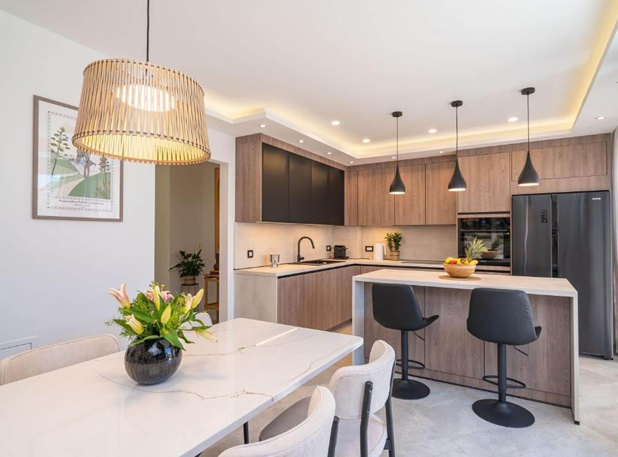 A modern kitchen with wooden cabinets, a central island, pendant lighting, and a dining area with a vase of flowers.