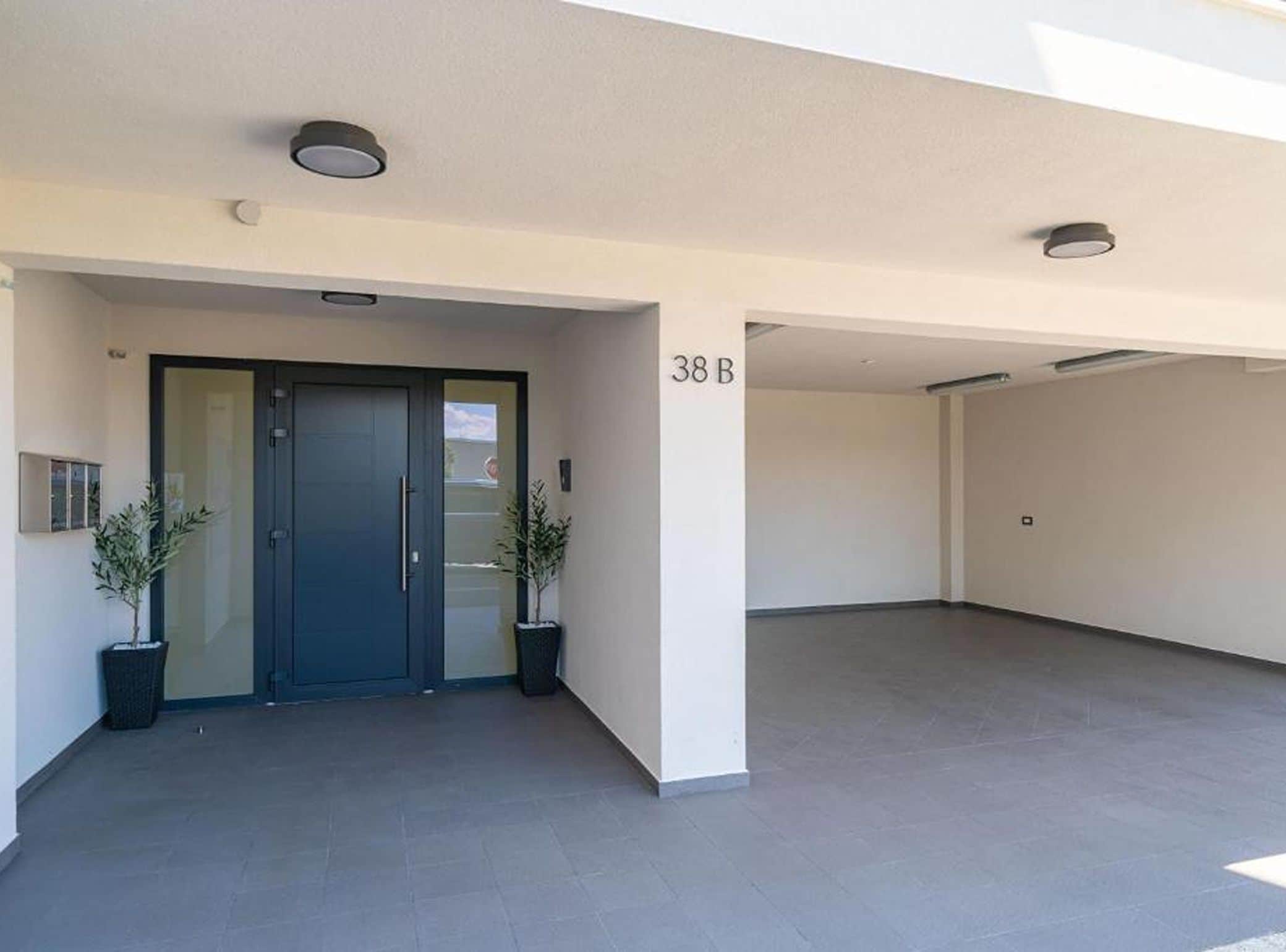 Entrada de un edificio con puerta oscura, plantas en macetas y techo plano.