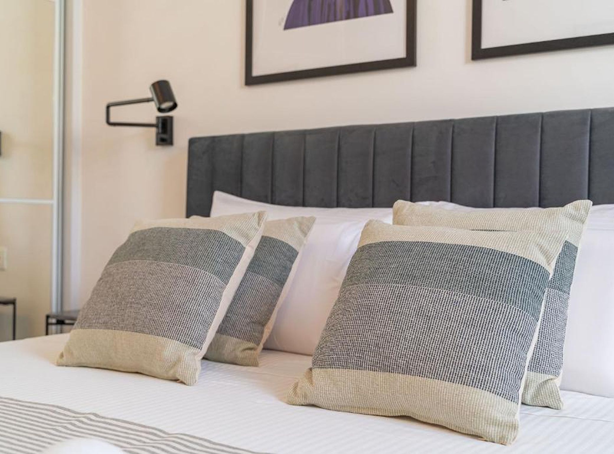 A hotel room with a bed, pillows, a headboard, a nightstand, and a wall-mounted lamp.