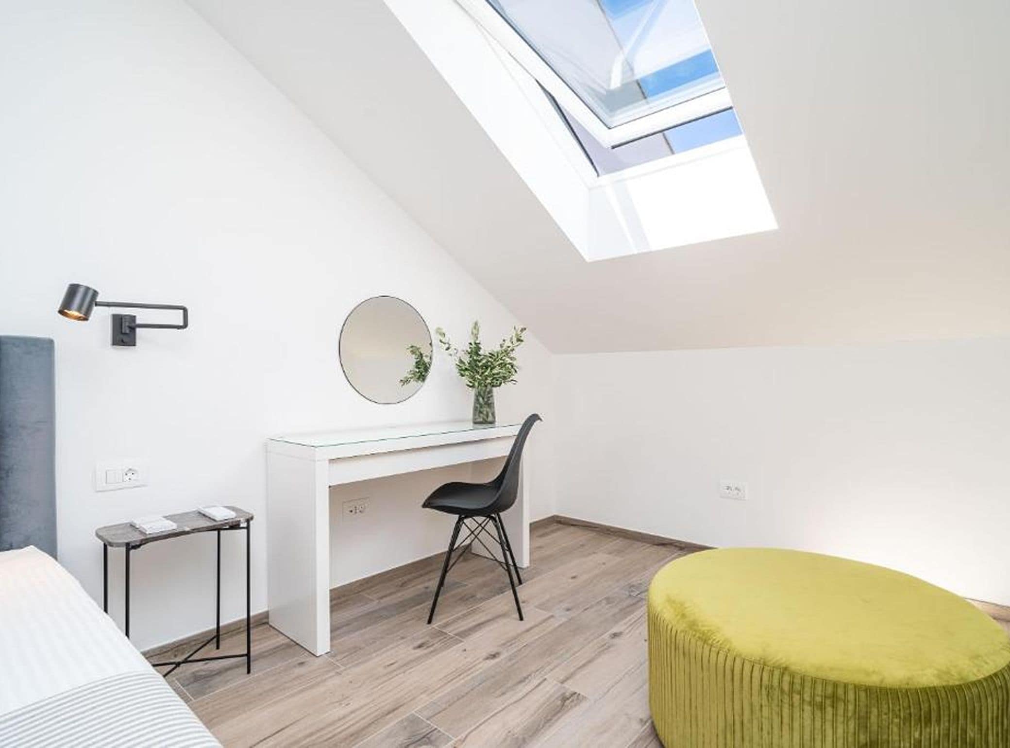 A modern loft bedroom with a skylight, a white desk, a black chair, and a green ottoman.