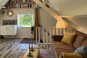 A loft-style room with a sloped ceiling, a white crib, a brown sofa with cushions, a patterned rug, a bookshelf, and a window with curtains. - Ouddorp Vakantiehuisje Zon en Zee (OUDDORP ZH)