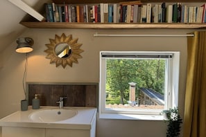 A bathroom with a sink, a mirror, a shelf with books, and a window with a view of greenery. - Ouddorp Vakantiehuisje Zon en Zee (OUDDORP ZH)