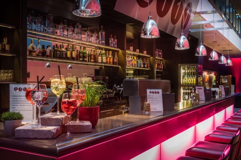 Una zona de bar con estanterías llenas de botellas, una barra con vasos y un cartel con un número.