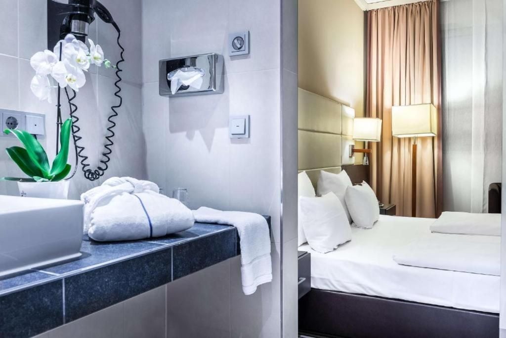 Una habitación de hotel con baño, una cama con ropa de cama blanca y una mesita de noche con lámpara.