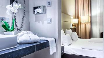 Una habitación de hotel con baño, una cama con ropa de cama blanca y una mesita de noche con lámpara.