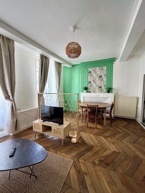 A room with a television on a wooden stand, a dining table with chairs, a patterned wall, and a radiator. - L'émeraude - Charming Apartment in Riom (Riom)