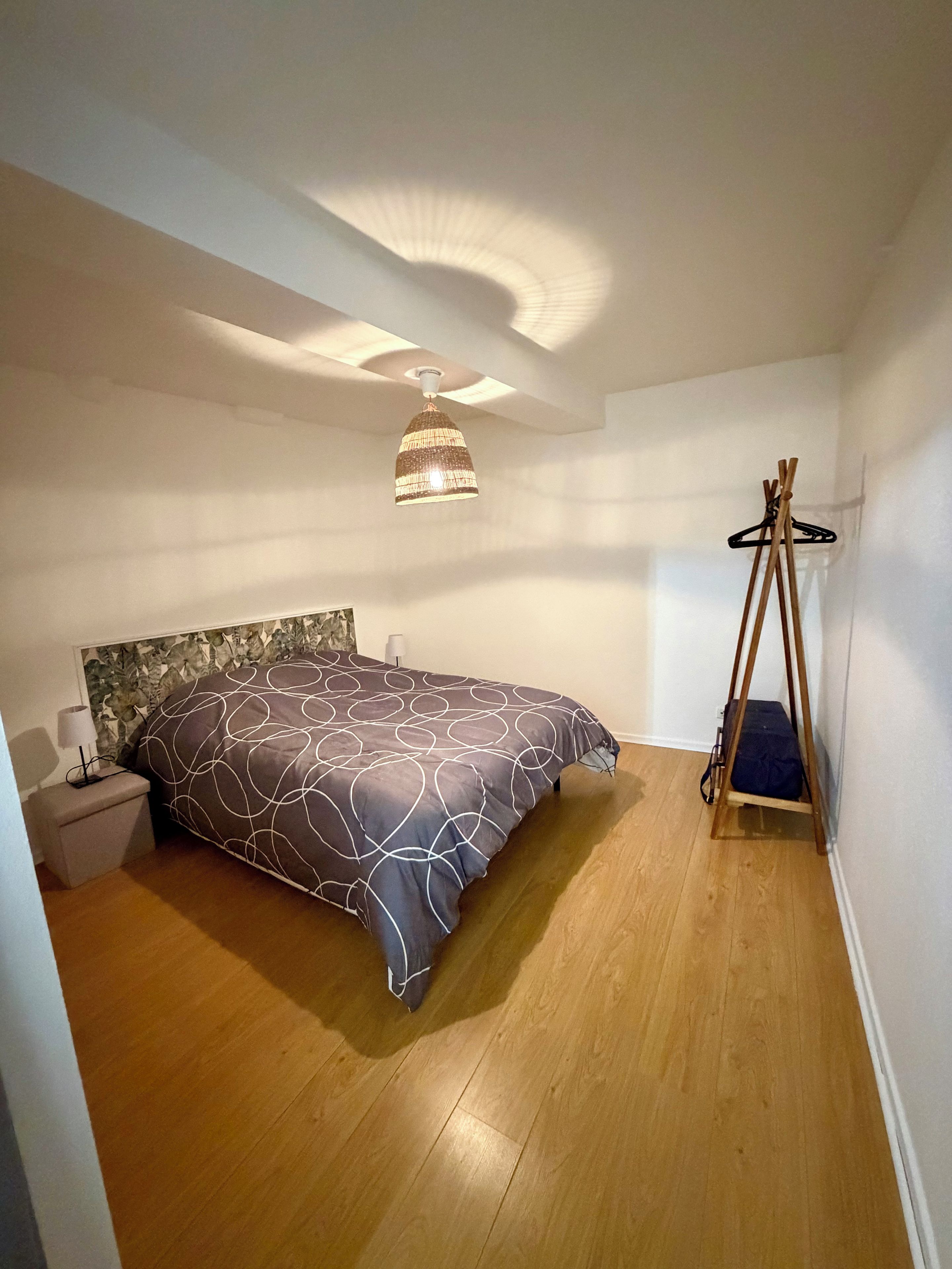 Quarto com cama, luminária pendente, mesa de cabeceira e cabide de madeira.