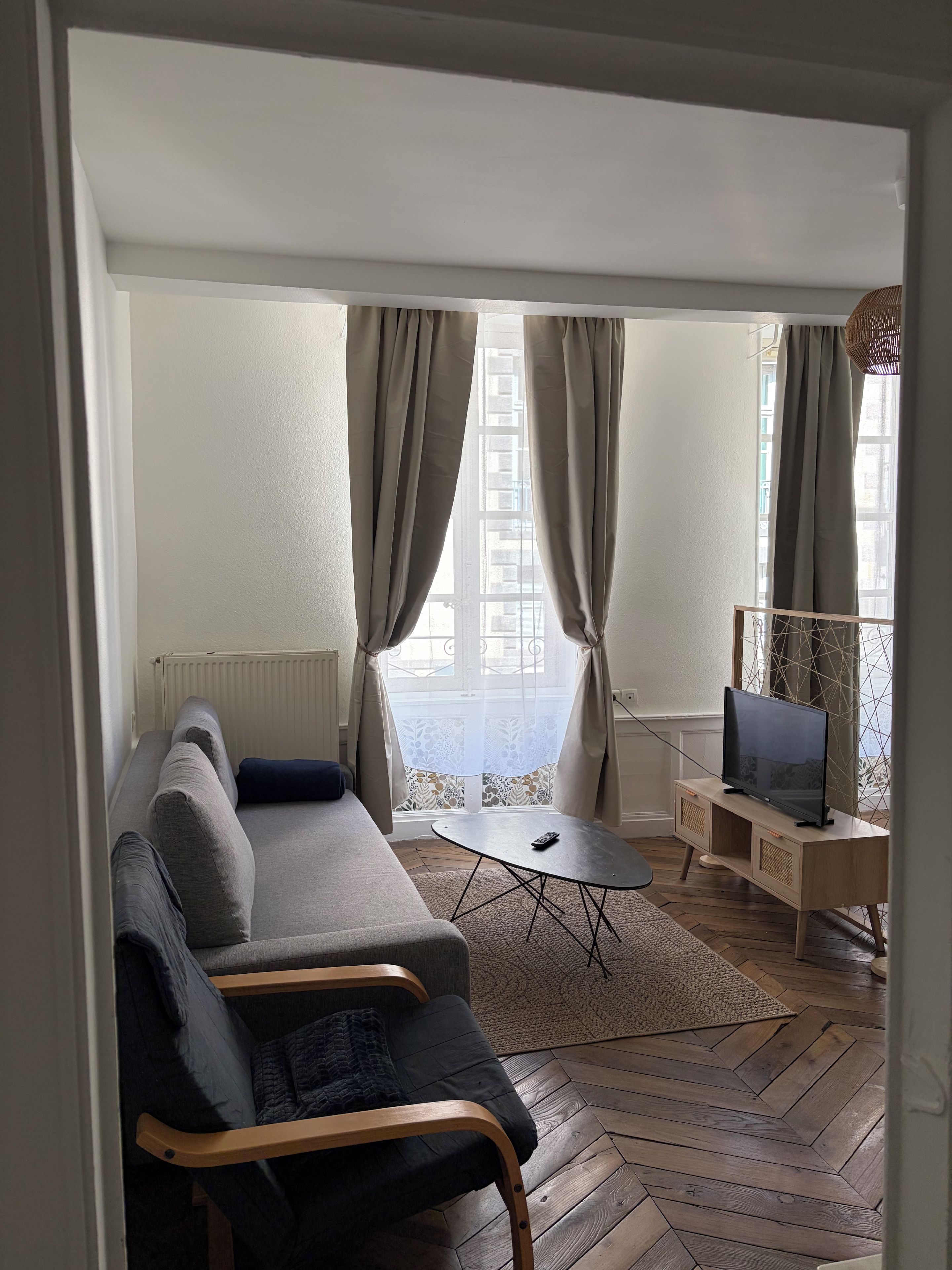 Ein Wohnzimmer mit einer Couch, einem Fernseher auf einem Schrank und Blick durch ein Fenster mit Vorhängen.