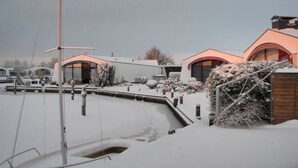 Área externa coberta de neve com um cais, um edifício com telhado curvo e um arbusto com neve.