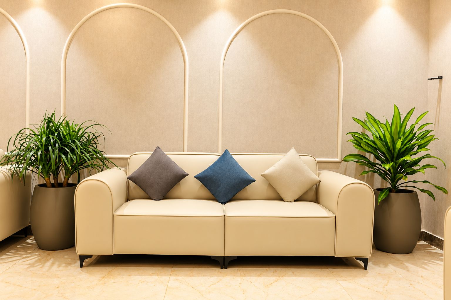 A beige sofa with three cushions, flanked by two potted plants, in a room with a marble floor and a wall with three arched niches.