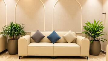 A beige sofa with three cushions, flanked by two potted plants, in a room with a marble floor and a wall with three arched niches.
