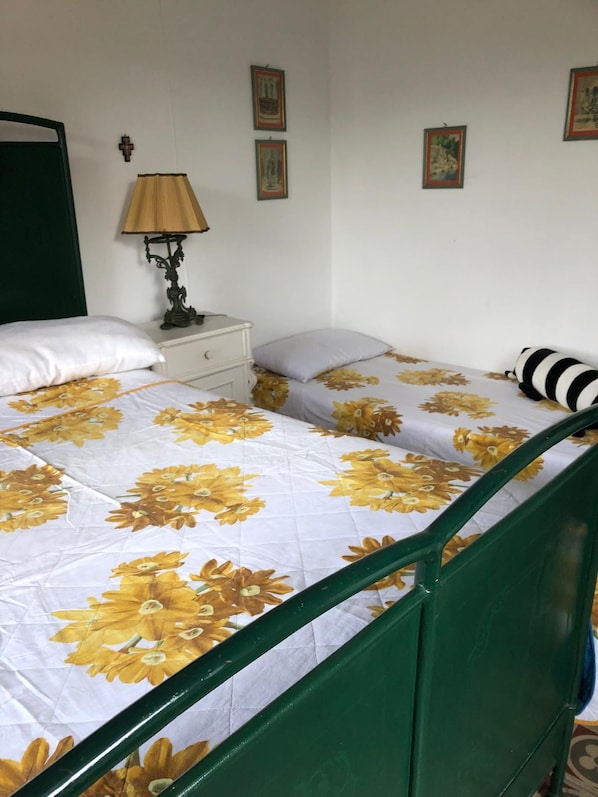 A single bed with a floral bedspread, a bedside table with a lamp, a nightstand, and a wall-mounted cross. - Vrbo Property (Liguria)