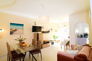 Ein modernes Wohnzimmer mit einem Glastisch, einer Couch, einem Fernseher und einer Küchenzeile im Hintergrund.