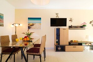 Ein Essbereich mit einem Glastisch, gepolsterten Stühlen, einem flachbildfernseher an der Wand und einem Regal mit Dekorationen.