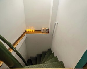 A staircase with a wooden handrail and a metal banister, leading to a landing with a shelf holding books and candles.