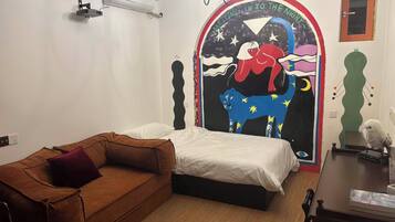 Un dormitorio con cama, un sofá marrón, unidad de aire acondicionado, candelabro y pared con un mural colorido.